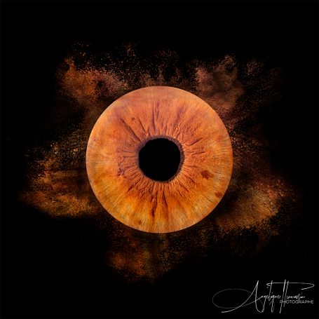 Iris Un œil orange sur fond noir avec des teintes de poussière colorée autour.